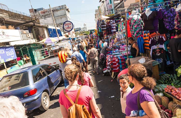 Street market in San Salvador. Long line of market stalls, cars on the street and people, tourists, walking along the street