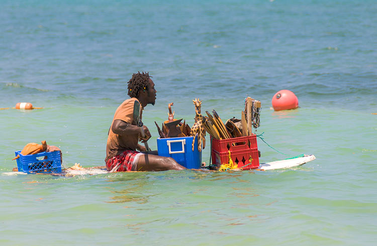 A Jamaican beach trader takes his goods afloat and sells souvenirs to tourists on the beach from a surf board