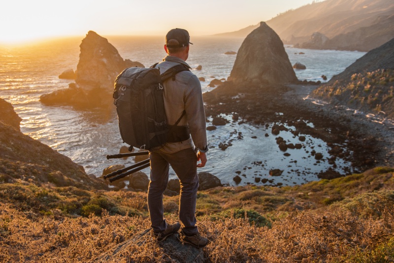 A man standing on a cliff side staring at the ocean wearing a backpack