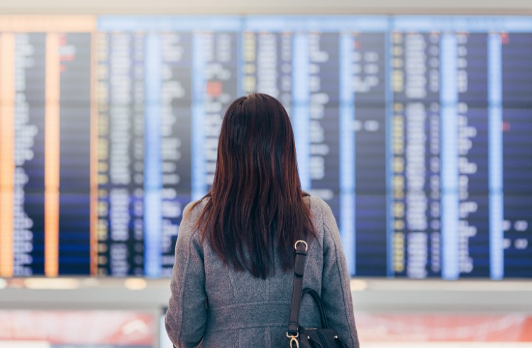 A woman stands in front of a flight information display at an airport.
