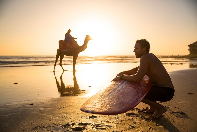 A surfer on the sand resting his board on his legds, with a person riding a camel visible in the background