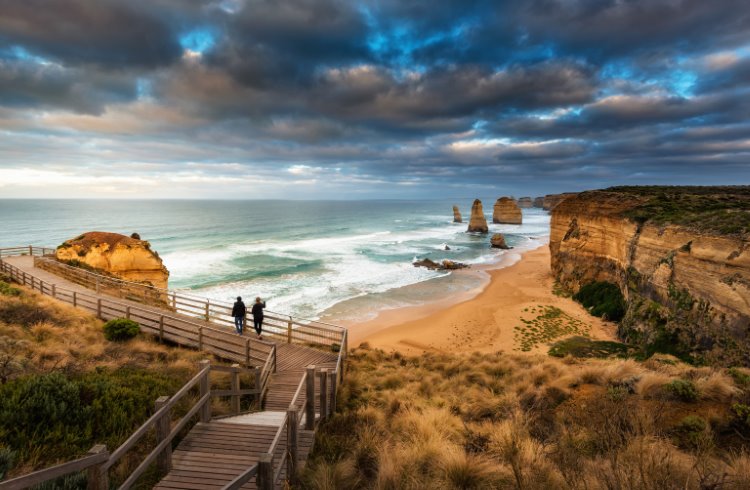 Two people look down from a seaside boardwalk at the 12 Apostles rock formations in Victoria, Australia. 