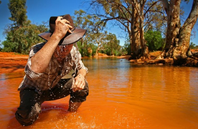 A man crouches beside a river in the Australian outback, pouring water from his hat onto his head.