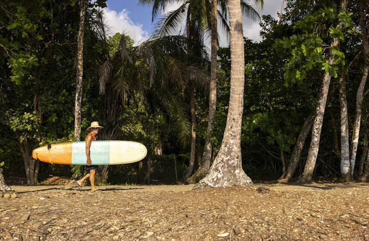 An older man carries a surfboard down a beach in Costa Rica.