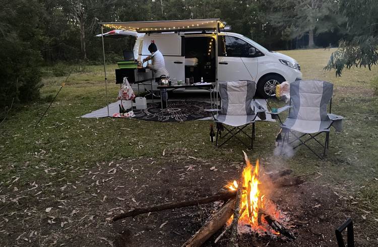 A woman cooks over a stove near her campervan in New South Wales, Australia, with a campfire in the foreground. 