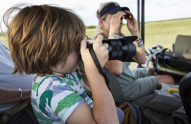 A young boy photographs wildlife from an open jeep on safari in Africa.