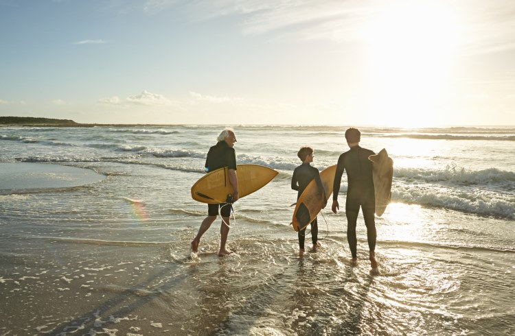 A boy, his father, and his grandfather carry surfboards on a beach.