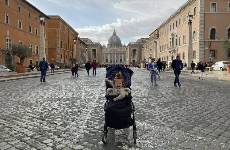 In the middle of a cobbled street in Vatican City, Italy, a baby in stroller smiles at the camera.