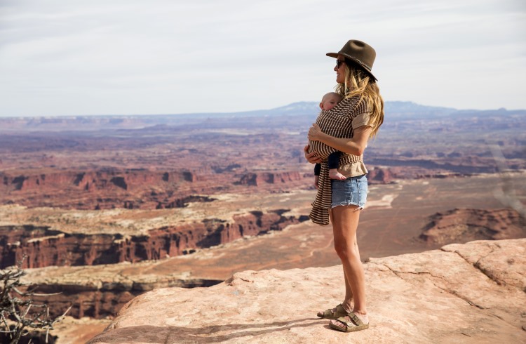 A woman with a baby in a sling across her chest stands at the edge of a canyon in Utah.