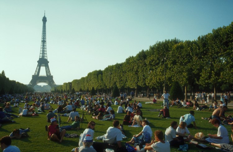 Visitors sit in a grassy park in Paris with the Eiffel Tower in the background. 