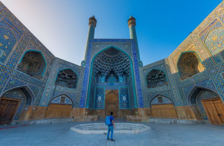 A male backpacker stands in front of the ornate Shah Mosque in Isfahan, Iran.