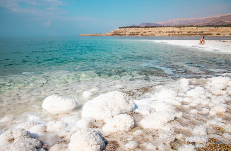 A woman sits at the edge of the Dead Sea in Jordan, surrounded by salt formations.