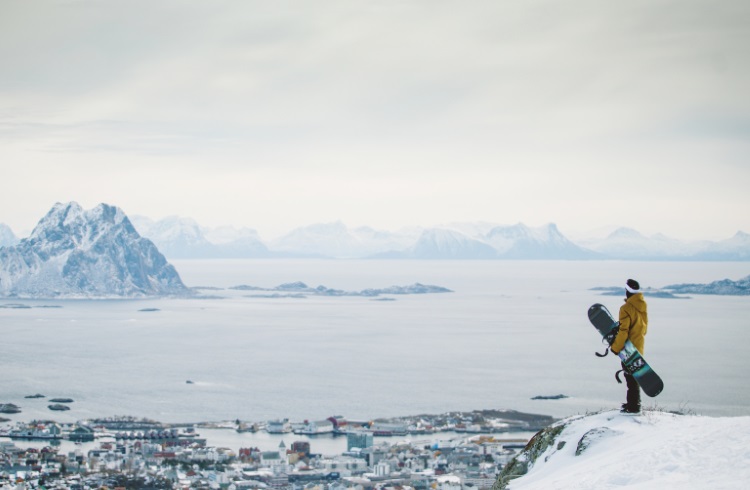 A snowboarder looks down over the sea from a snowy peak in the Lofton Islands, Norway.