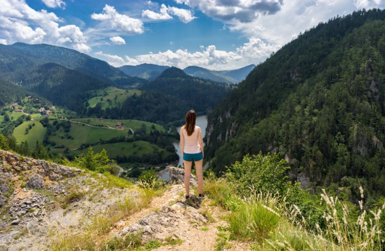 A young female hiker stands overlooking a lake and green, wooded hills in Tara National Park, Serbia.