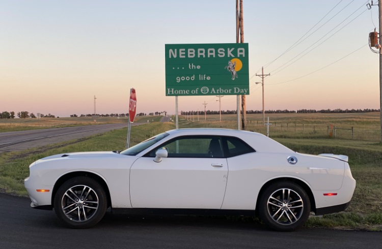 A white Mustang car parked by the highway, in front of the Nebraska welcome sign at the state line.