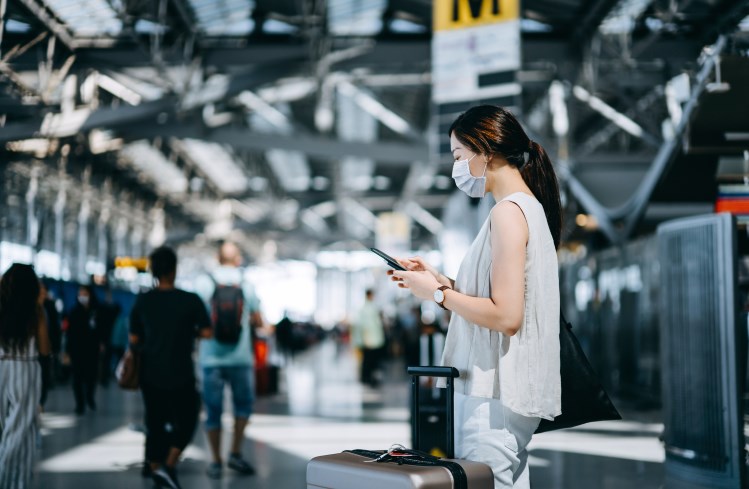 A woman in a face mask stands in the Bangkok airport, checking flight information on her smartphone.