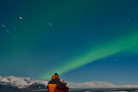 A couple with their arms around each other, standing in a snowy landscape watching the northern lights