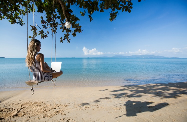 A woman sitting on a swing on the beach while using her laptop