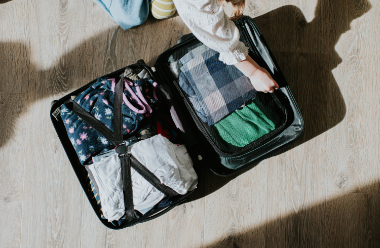 Top down conceptual image of a child kneeling on the floor. She opens a neatly packed suitcase, filled with clean, folded clothing.