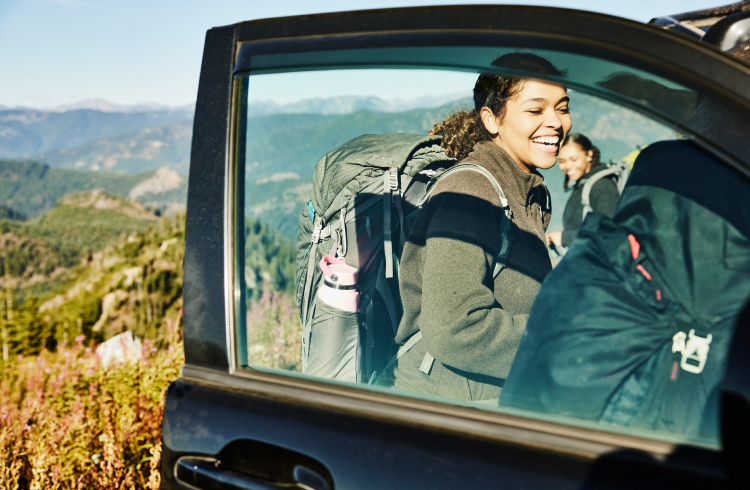 A laughing young woman wearing a backpack prepares for road trip with sisters