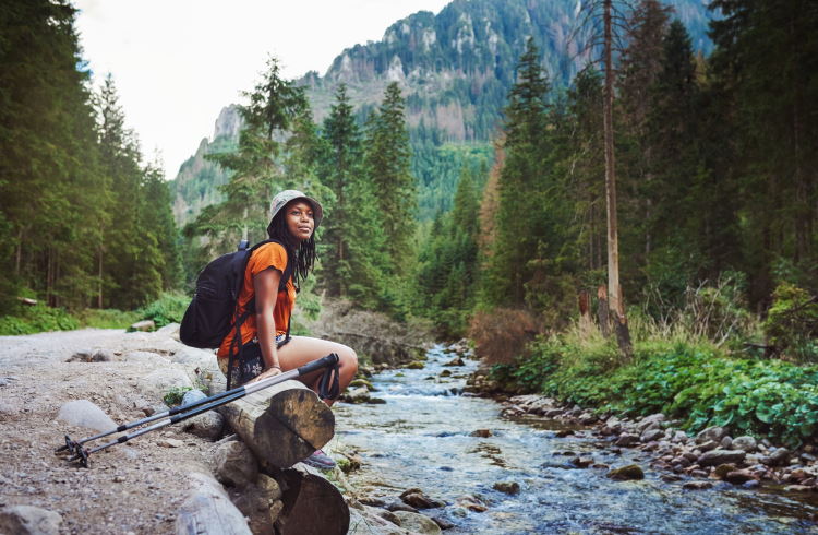 A woman hiker sits beside a stream, with hiking poles laid next to her.