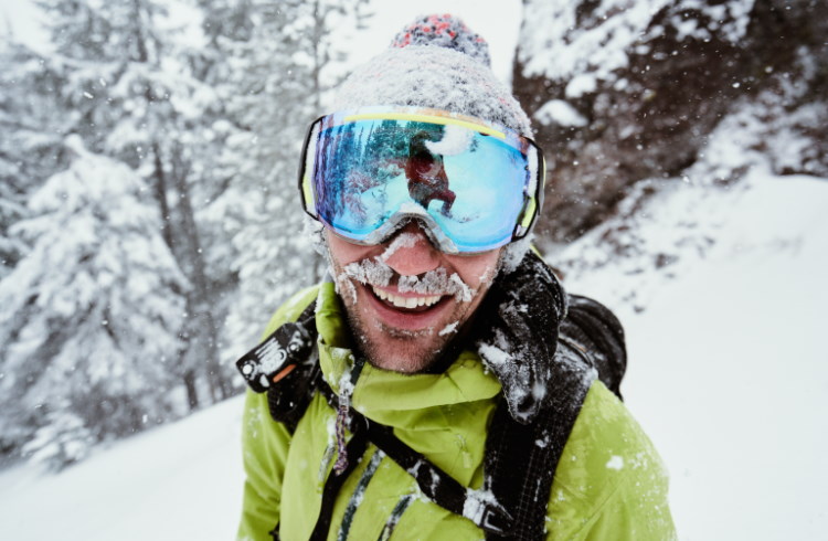 A goggle-wearing skier covered in snow smiles cheerfully at the camera.