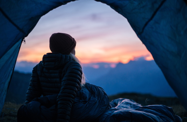 A woman in a down jacket looks out from her camping tent in the mountains.