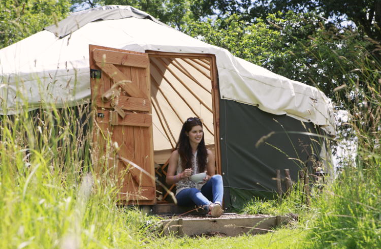 A woman sits in the doorway of a yurt out in the wilderness.