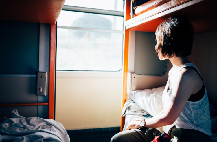 A woman sits on a bunk bed and gazes out the window of a sleeper train car.