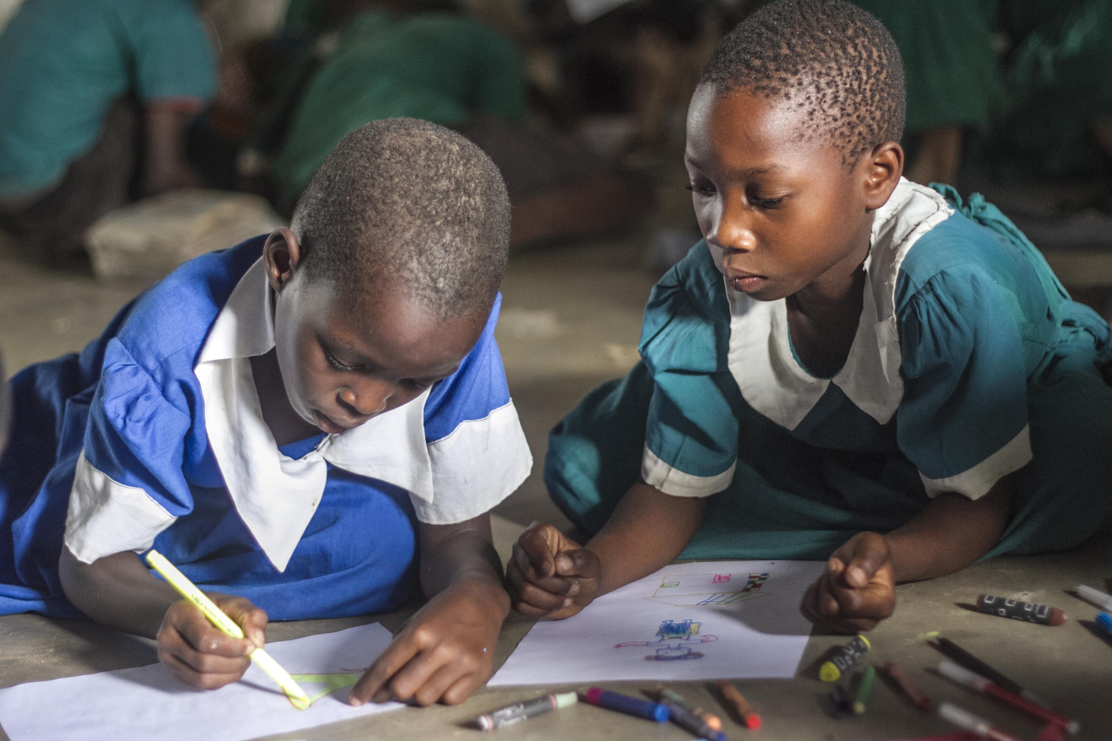 Two girls from Standard Two using coloured pens and pencils for the first time.  Quite often class members need to share a pencil between two. The students sit on the concrete floor, as there are no desks or chairs at the school. 