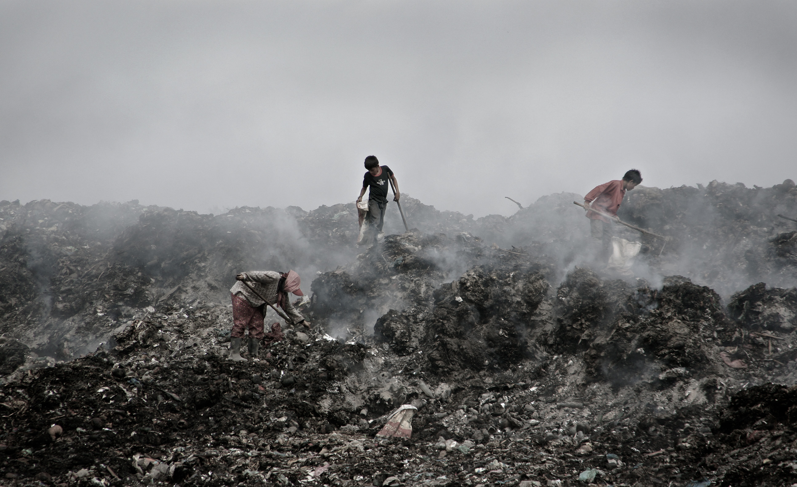 Workers walk across a smoky, smoldering mountain of waste in search for any last items that have been left behind that can still be salvaged for recycling. Any items found are put into their bags that are strapped around their waist.