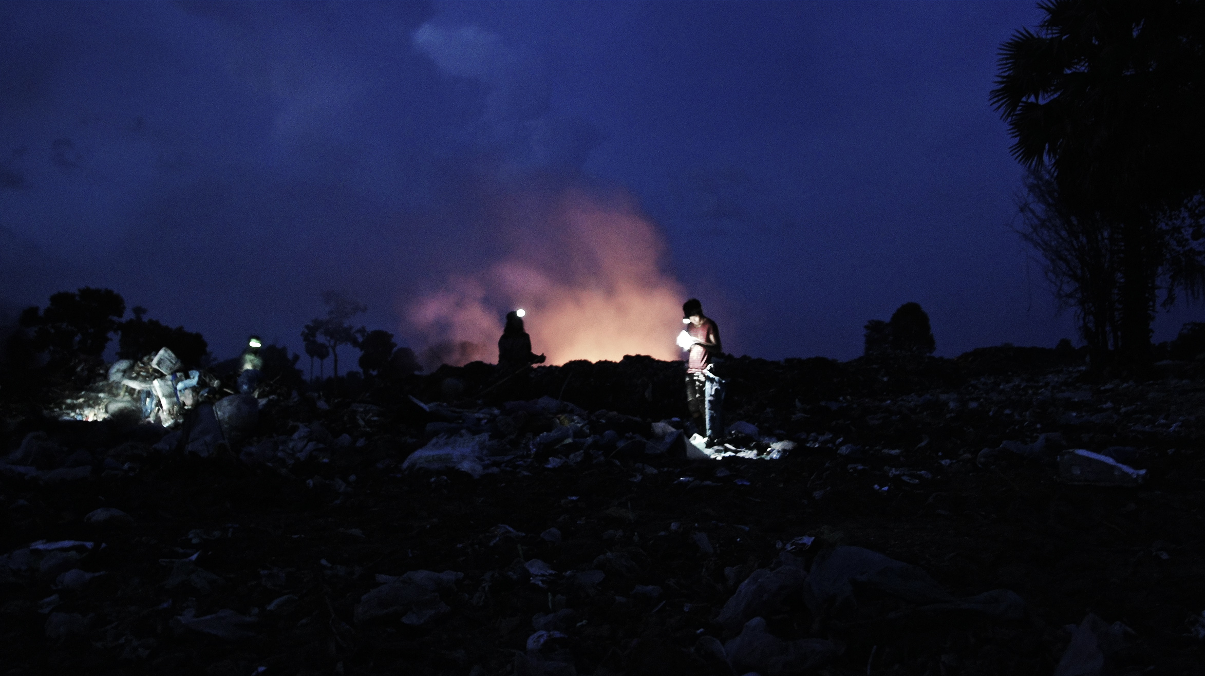 As darkness falls, work still continues late into the night; Sobat, a 17-year-old high school student by day straps on his head torch and searches for recyclable items. He comes to the dump at night to try to make enough money to send himself to college.