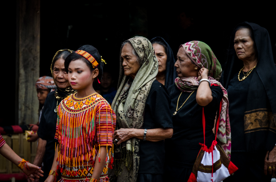 Three generations of women attend the funeral service of their family member. Men and women parade separately through the funeral ceremony numerous times over the course of the day. It is an occasion that unites the family and celebrates their whole ancestry.