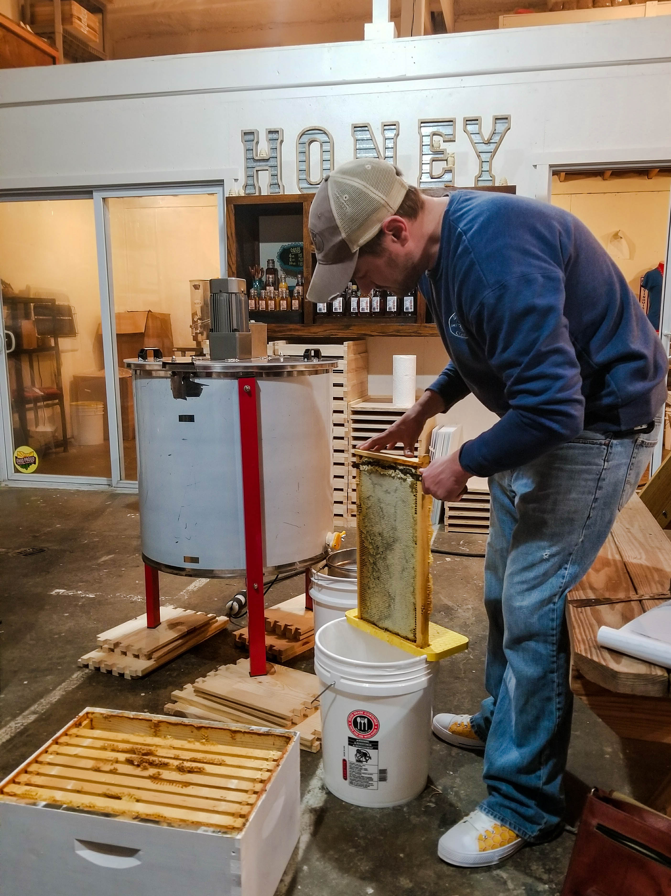 The honey harvest has begun. Here, the beekeeper uses a warm knife to remove the wax cappings that seal the honey in the comb. The frame will be inserted into the centrifuge behind him, and the honey will be flung out to drip down to the bottom where it will be filtered and strained into jars.
