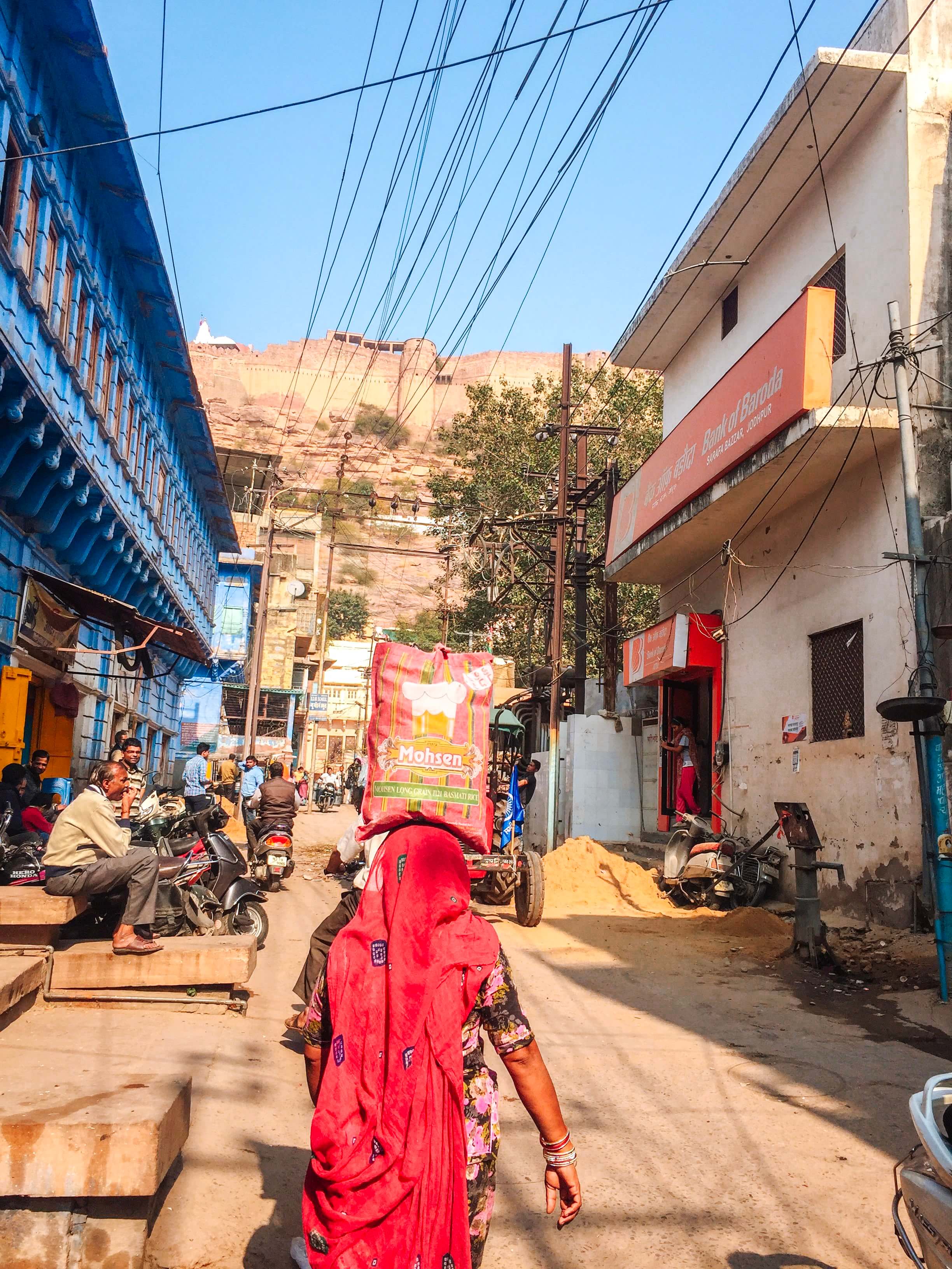 A woman in ruby expertly balancing rice on her head as she begins her ascent up the winding alleys and steeply inclined steps of the Blue City.