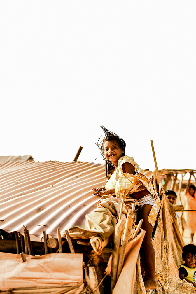 A young girl smiles for the camera after climbing up the side of her family's makeshift shed. Structures of this nature are common in the desert, as building materials are scarce and families must take advantage of what they have on hand to create shelter from the blistering sun. 