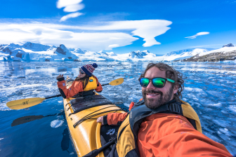 Jared and Alesha sit in a yellow kayak wearing orange jackets and yellow life vests. They look back and wave at a camera while paddling in icy water in antarctica