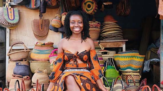 Tuli wears a brightly coloured dress while sitting and smiling in front of brightly coloured baskets