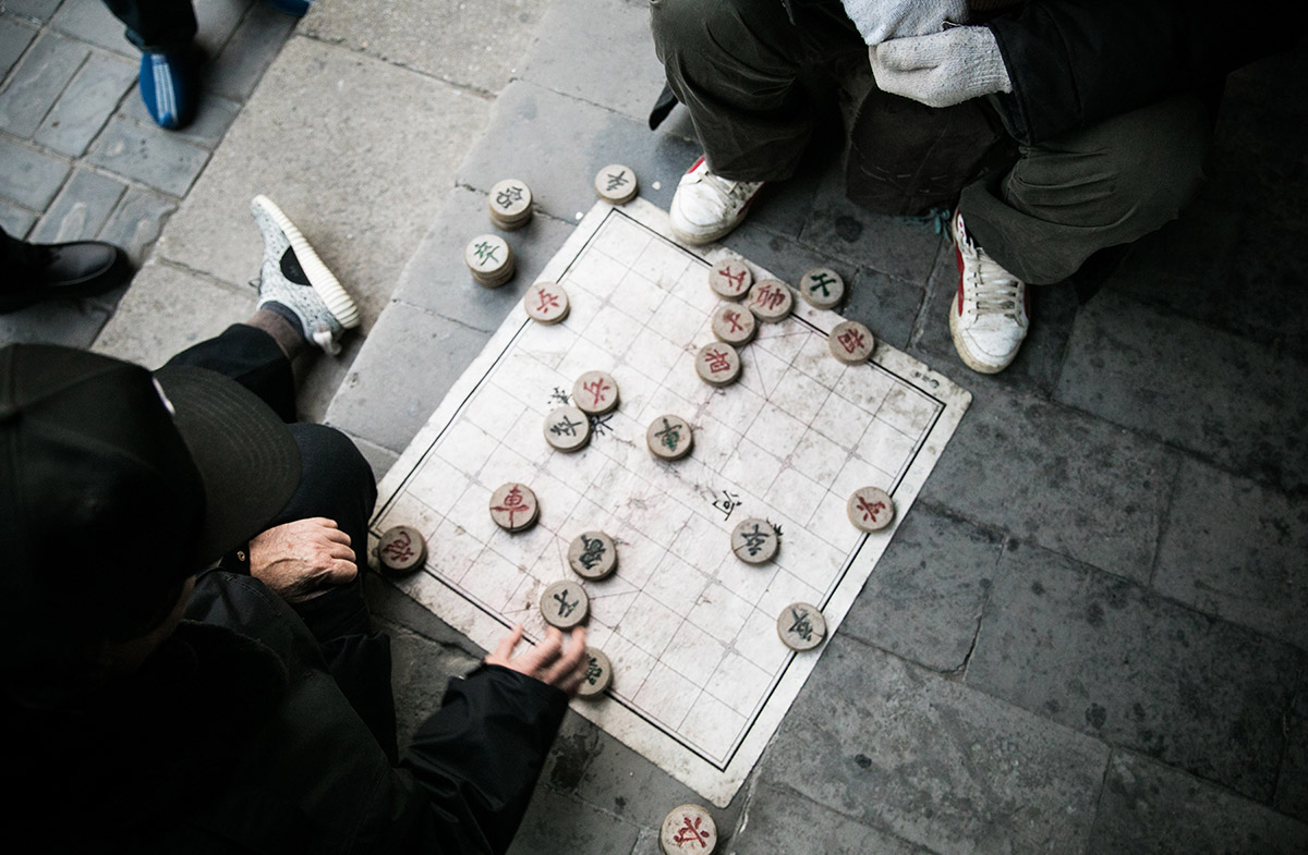 Two men play a game of "Xiangqi" on the Temple grounds – literally placing their playing mat on the concrete. Spectators watch eagerly over their game play, with the low angle of the board giving them the best view.