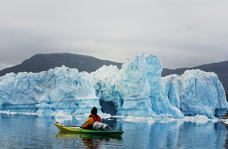 A person kayaking in Greenland near a large iceberg on a cloudy day