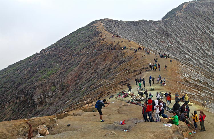 Crowds of tourists looking into a volcanic crater from a rocky mountain shrouded by clouds