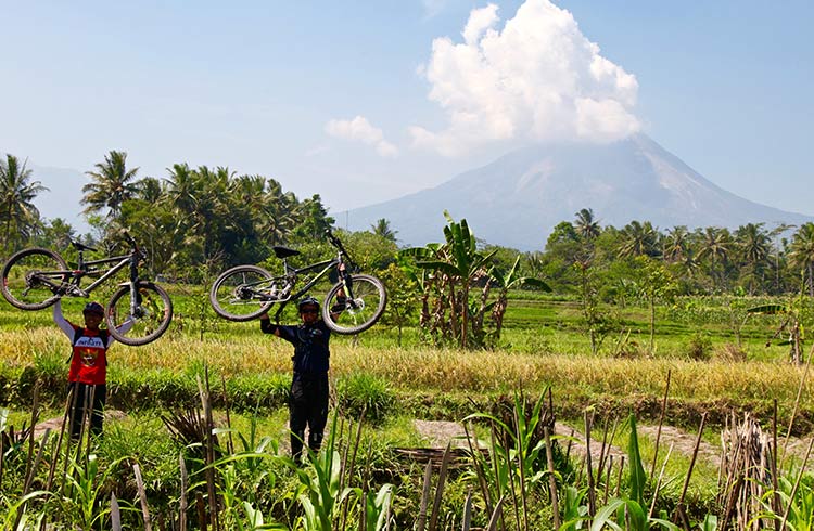 A volcano erupts in the background as mountain bikers in a grass paddock begin to leave the scene