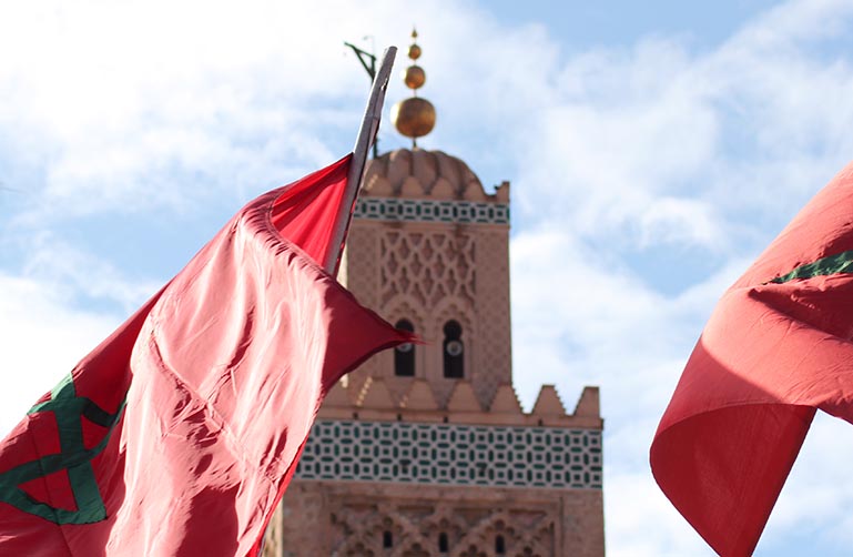 Moroccan flags wave in front of a minaret in Morocco.