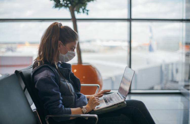 Une femme est assise dans une zone frontalière de l'aéroport portant un masque et à l'aide d'un ordinateur portable.