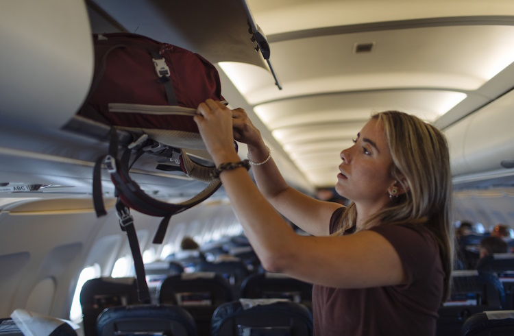A woman removes her carry-on bag from the overhead compartment of an airplane.