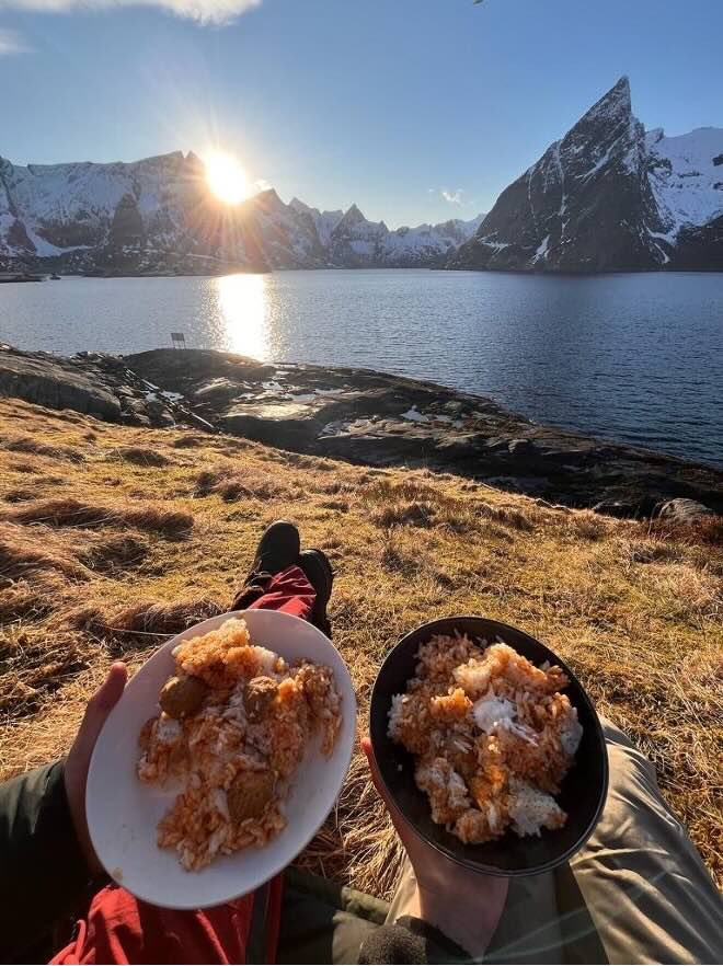 Two people holding their meals in front of a scenic mountain and lake