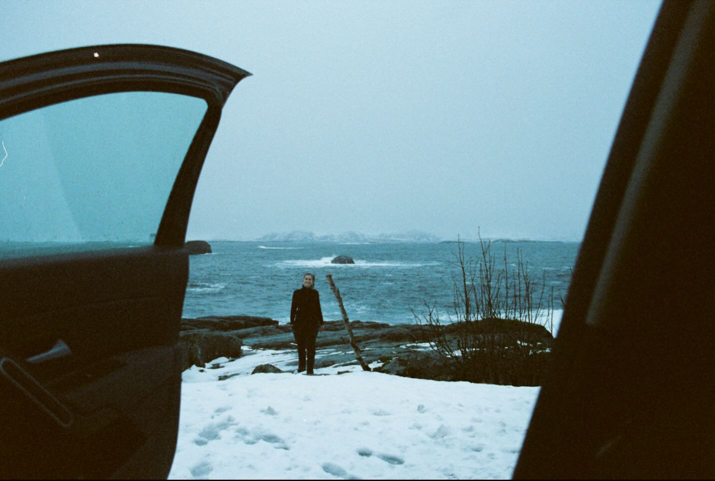 A woman standing in front of a frozen lake, photographed through the open door of a car