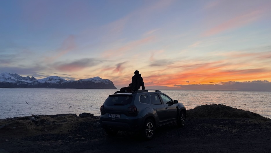 A person sitting on the roof of a car, in front of a lake at sunset