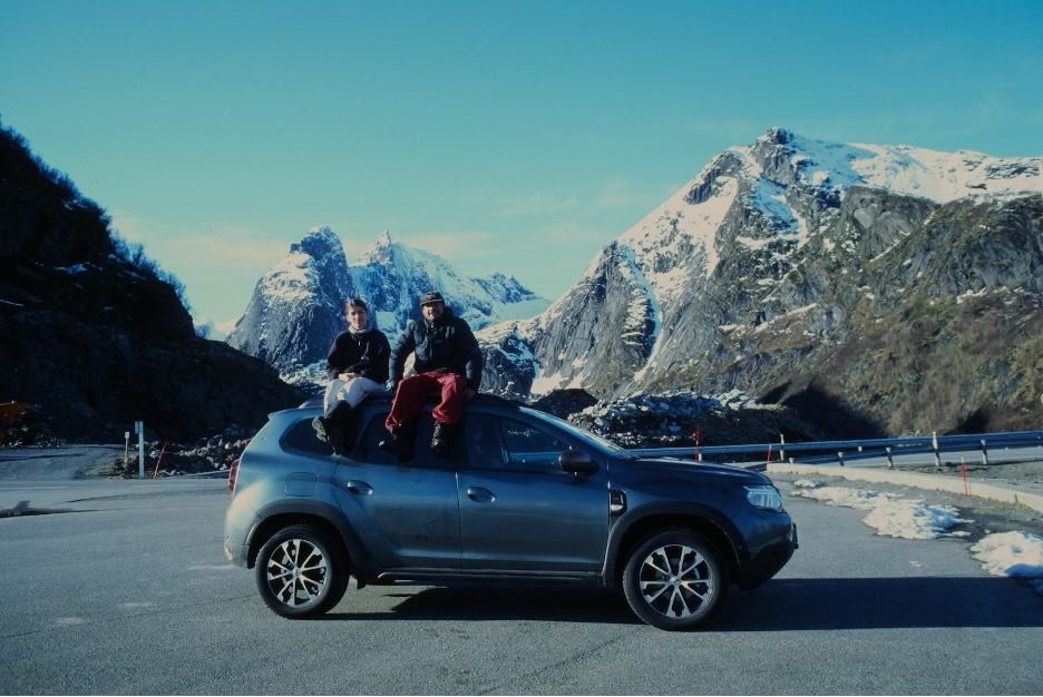 Two people sitting on top of a car roof in front of snow capped mountains