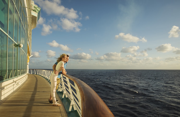 A young couple stands on the deck of a cruise ship, gazing out to sea. 